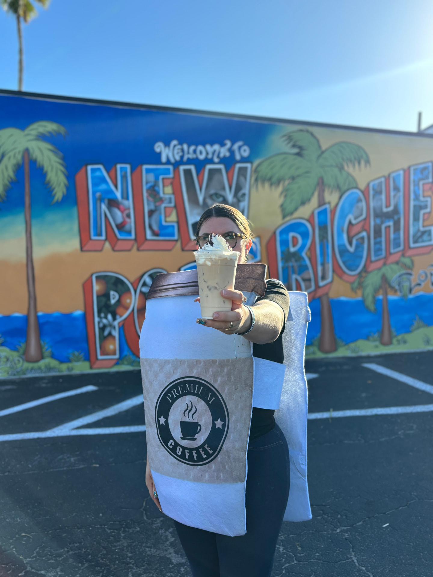 Sarah holding an iced latte in front of the Welcome to New Port Richey mural